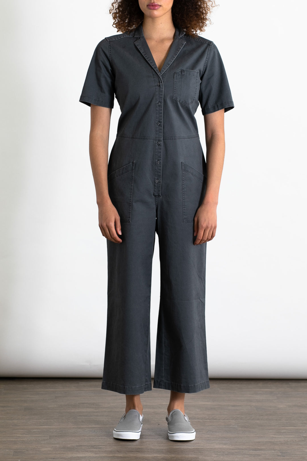A person stands against a plain background wearing the Bridge & Burn Lou Utility Jumpsuit in Slate, featuring a button-up front, side pockets, and adjustable buttons. They pair it with gray slip-on shoes and have their arms relaxed at their sides.
