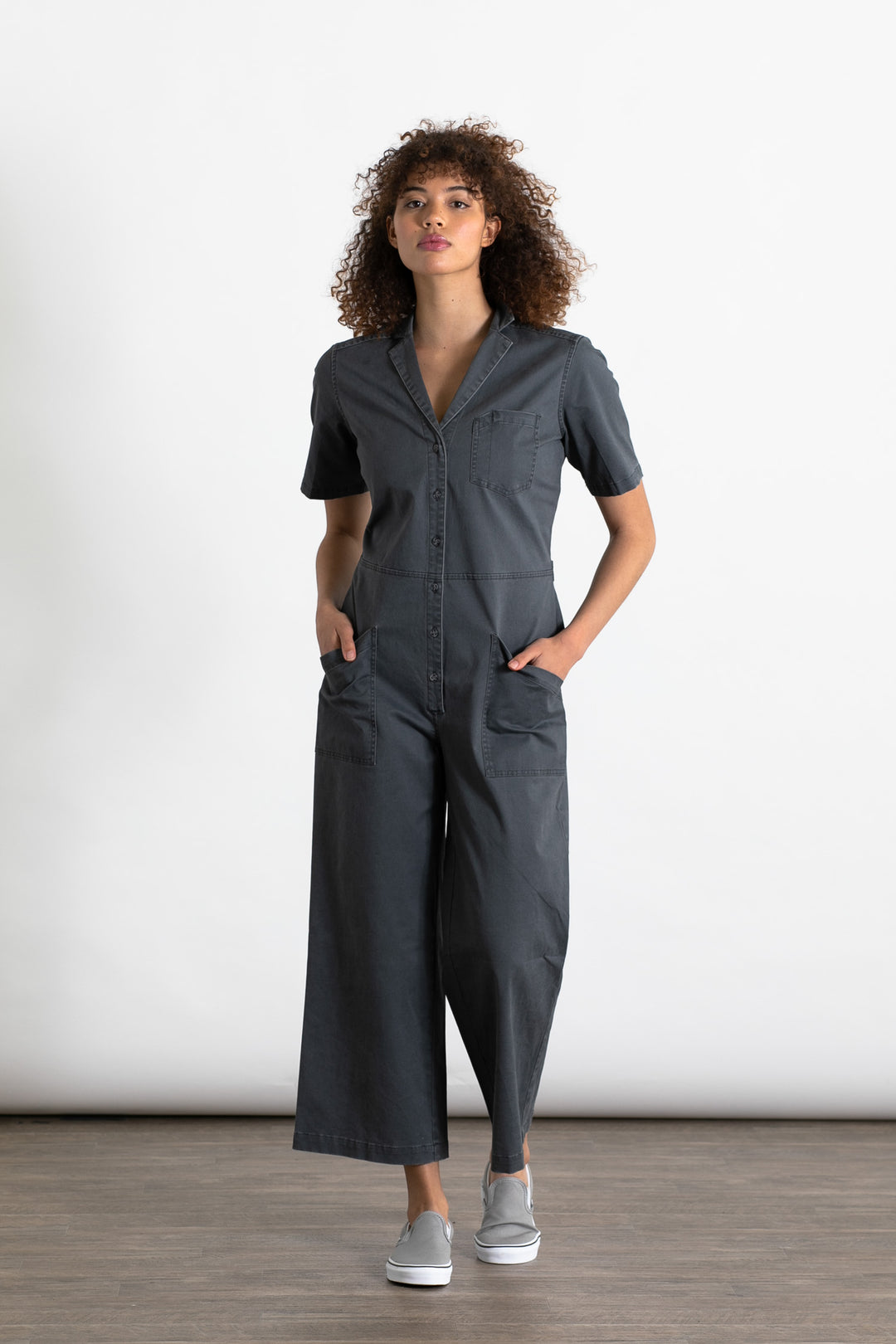 A person with curly hair wears the Bridge & Burn Lou Utility Jumpsuit in Slate, featuring adjustable buttons and front pockets, paired with gray slip-on shoes, standing against a plain white background with hands in pockets.