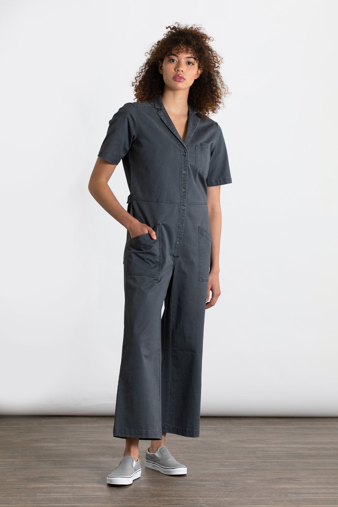 A person with curly hair wears the Bridge & Burn Lou Utility Jumpsuit in Slate—short-sleeve, wide-leg, adjustable buttons, and front pockets—with light gray slip-on shoes, standing indoors on a wooden floor against a white background.