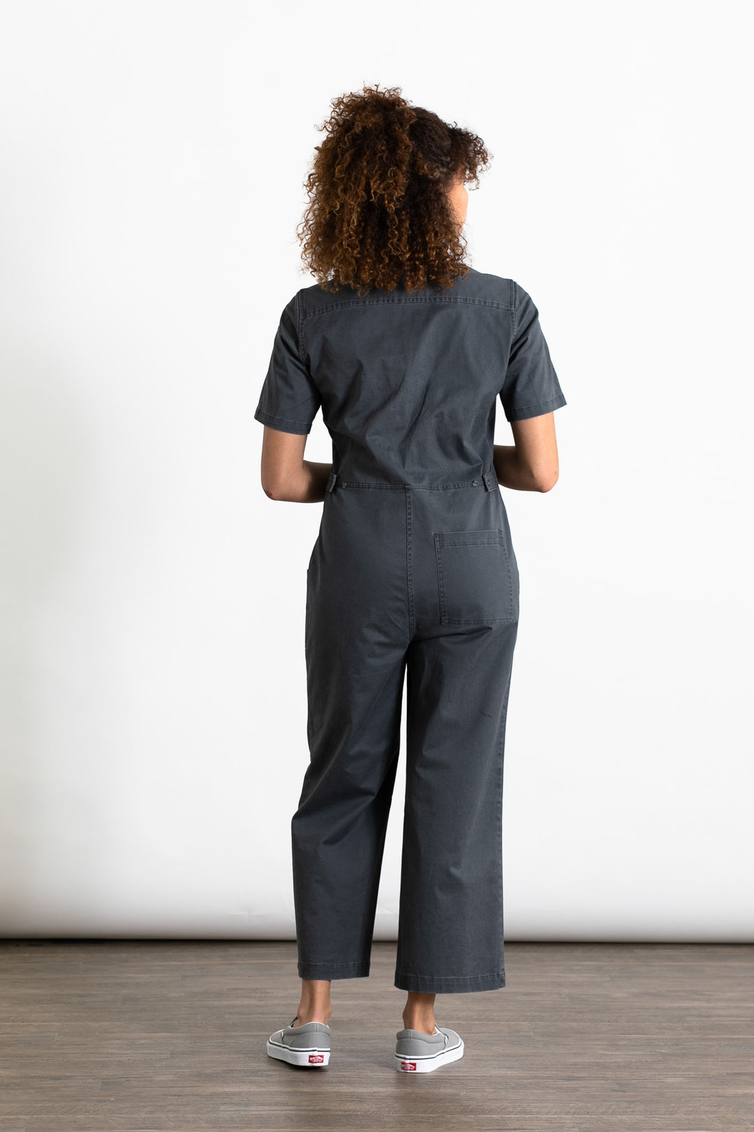 A person with curly hair, wearing the Bridge & Burn Lou Utility Jumpsuit in Slate and gray slip-on shoes, stands indoors facing away from the camera against a plain white background.