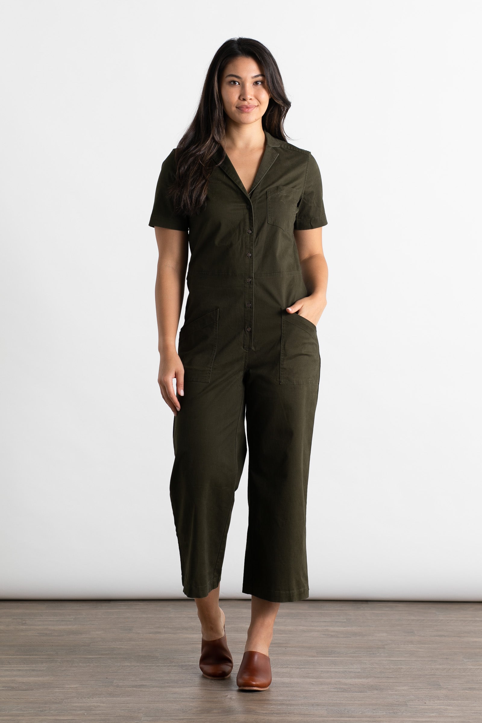 A woman with long dark hair stands on a wooden floor against a white background, wearing the Bridge & Burn Lou Utility Jumpsuit in Olive with short sleeves and brown shoes. Her hands are in her pockets.