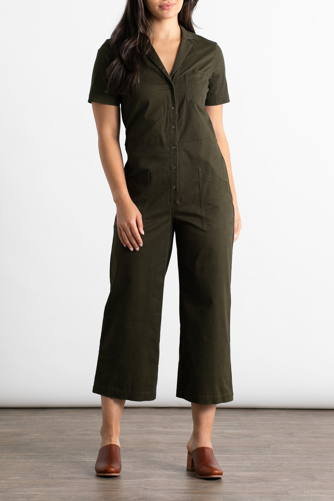 A woman stands on a wood floor wearing the Bridge & Burn Lou Utility Jumpsuit in olive, featuring an easy fit, button front, and wide cropped legs. She pairs it with brown clogs. Her head is partially out of the frame.