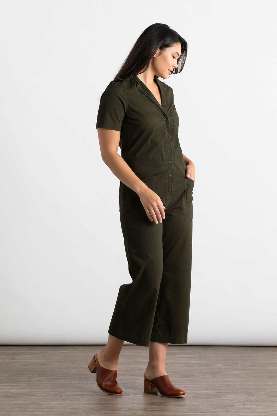 A woman with dark straight hair stands sideways against a white background, wearing the Bridge & Burn Lou Utility Jumpsuit in Olive with short sleeves and cropped wide legs, paired with brown heeled mules and one hand in her pocket.