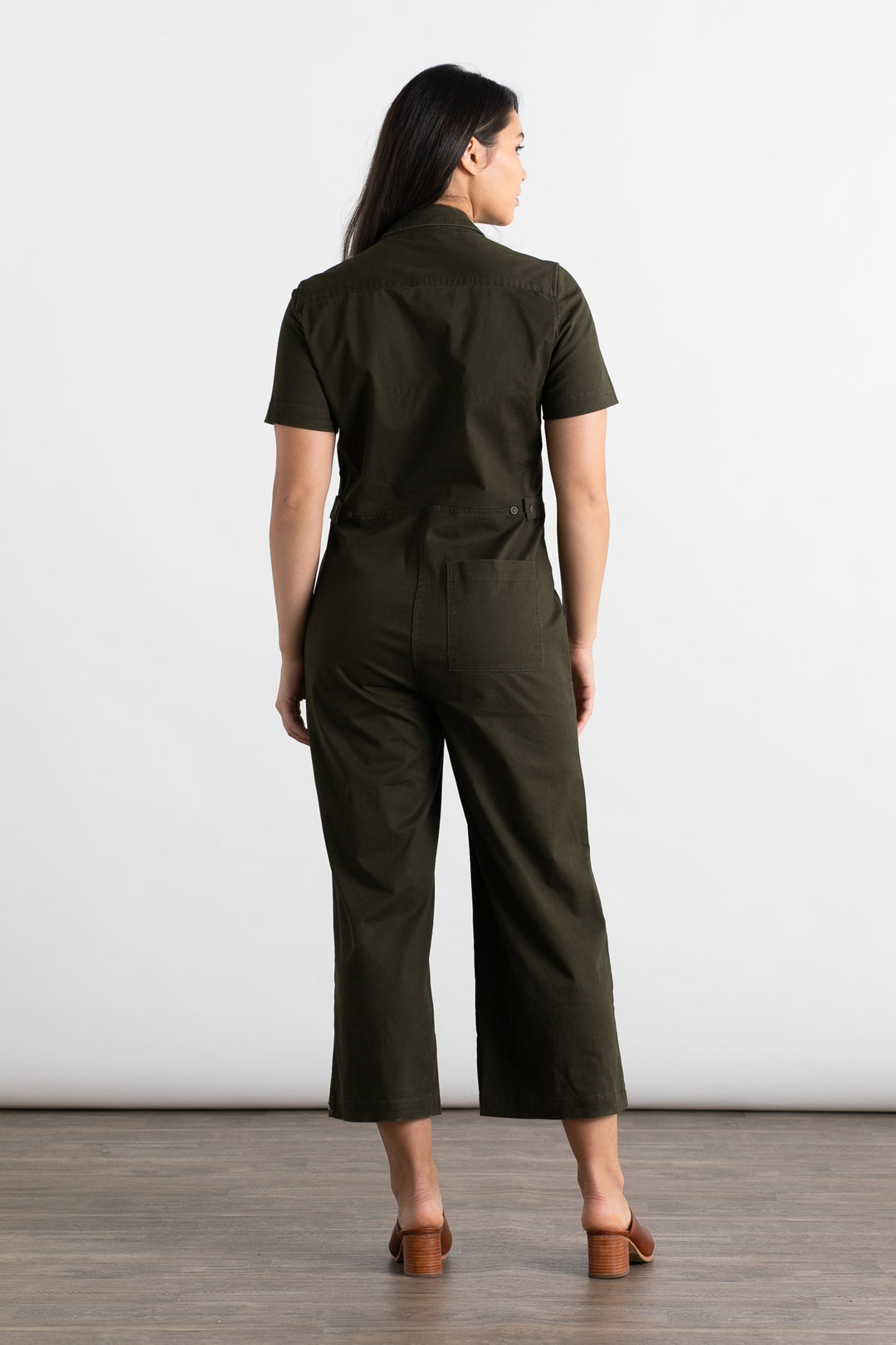 A woman faces away, wearing the Bridge & Burn Lou Utility Jumpsuit in olive with wide legs and brown block-heeled sandals, standing on a wooden floor against a plain white background.
