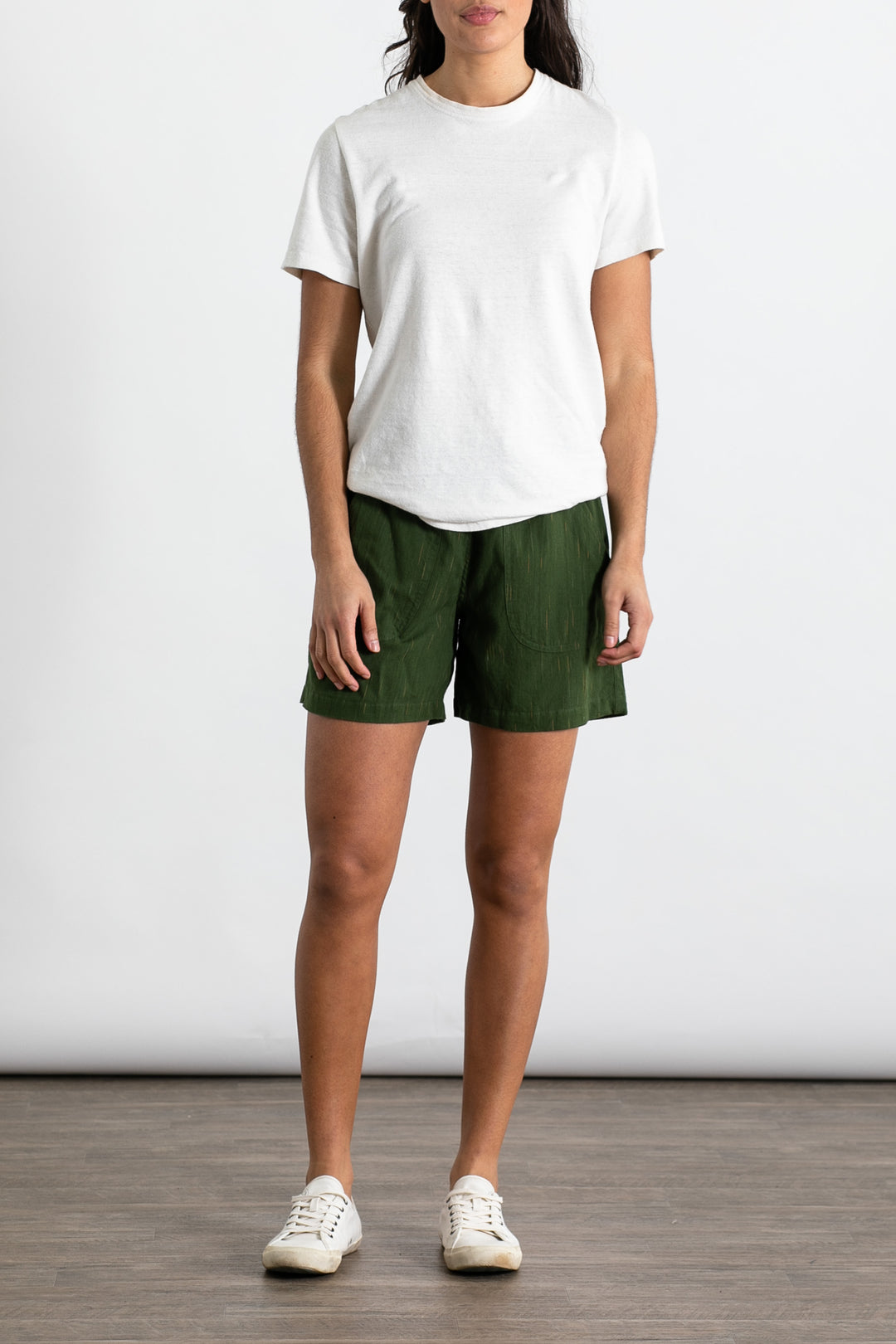 Indoors on a wooden floor, a person wears Jungmaven Baja Tee in Washed White with green shorts and white sneakers, arms relaxed by their sides, set against a plain light background.