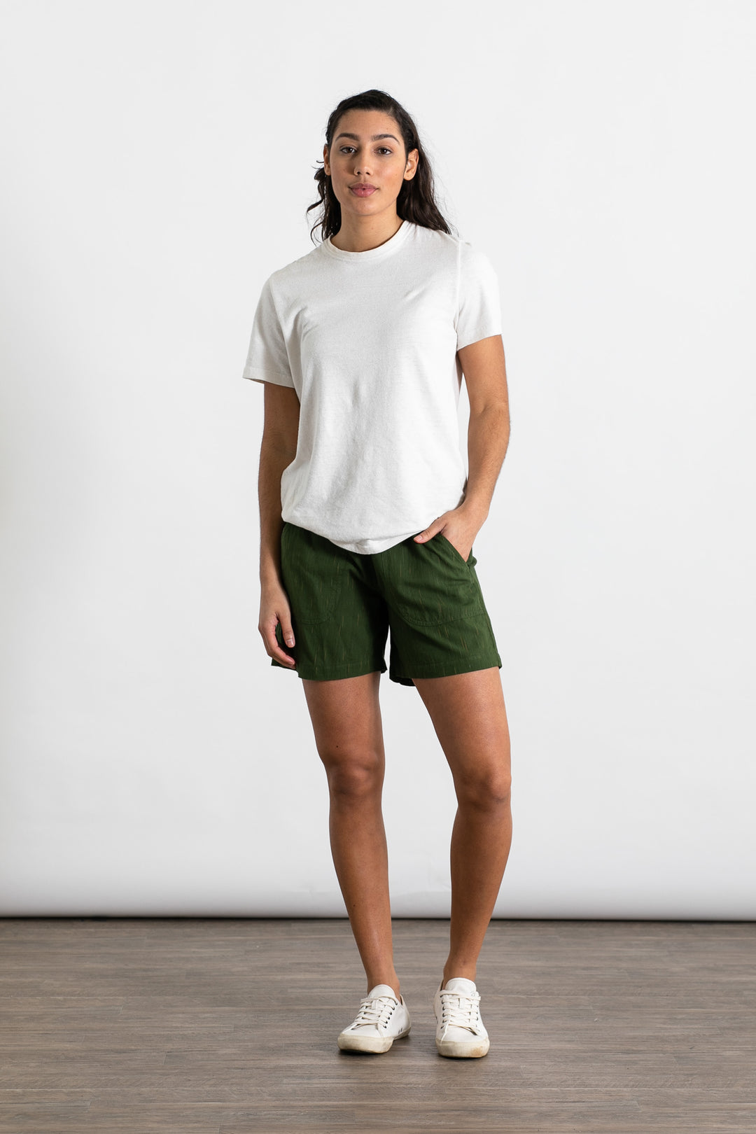 A person stands indoors against a plain white wall, wearing the Jungmaven Baja Tee in Washed White with dark green shorts and white sneakers, one hand in their pocket and a neutral expression.