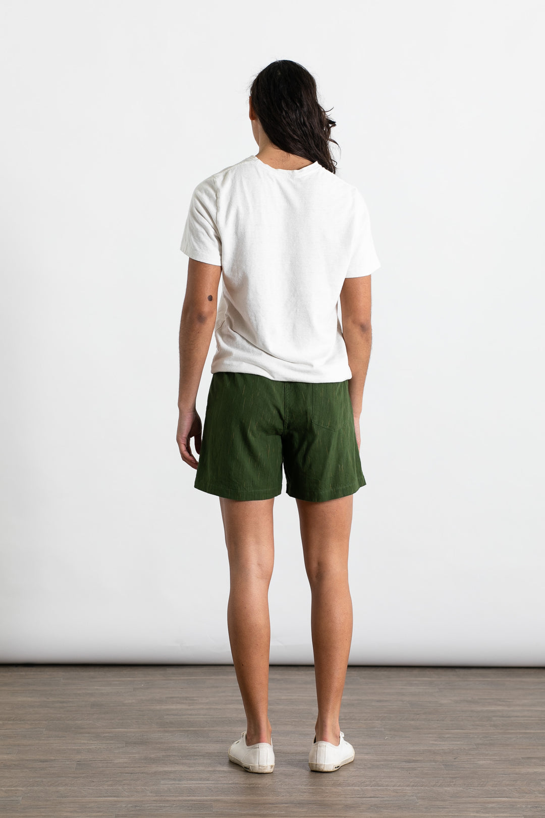 A person stands facing away from the camera, wearing the Jungmaven Baja Tee in Washed White and green shorts, with white slip-on shoes, against a plain white background with a wooden floor.