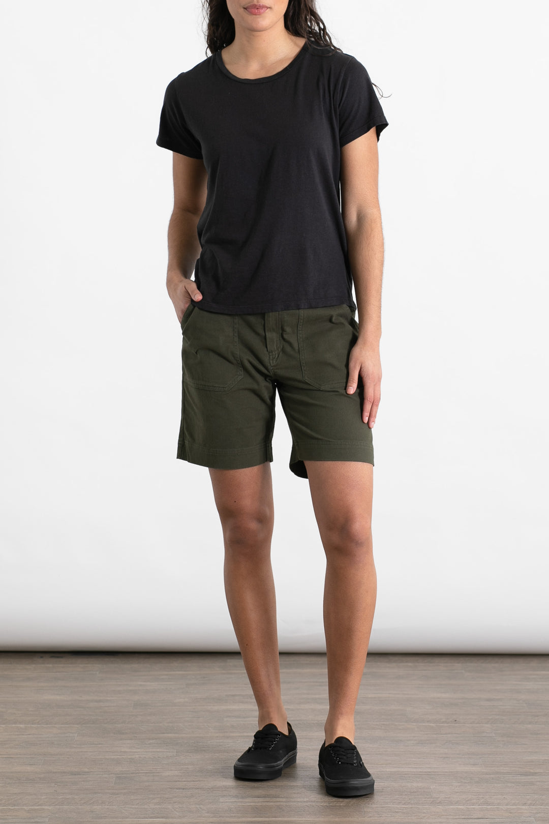A person stands against a plain background wearing a Jungmaven Ojai Tee in black, olive green shorts, and black sneakers, with one hand in their pocket and the other relaxed by their side.