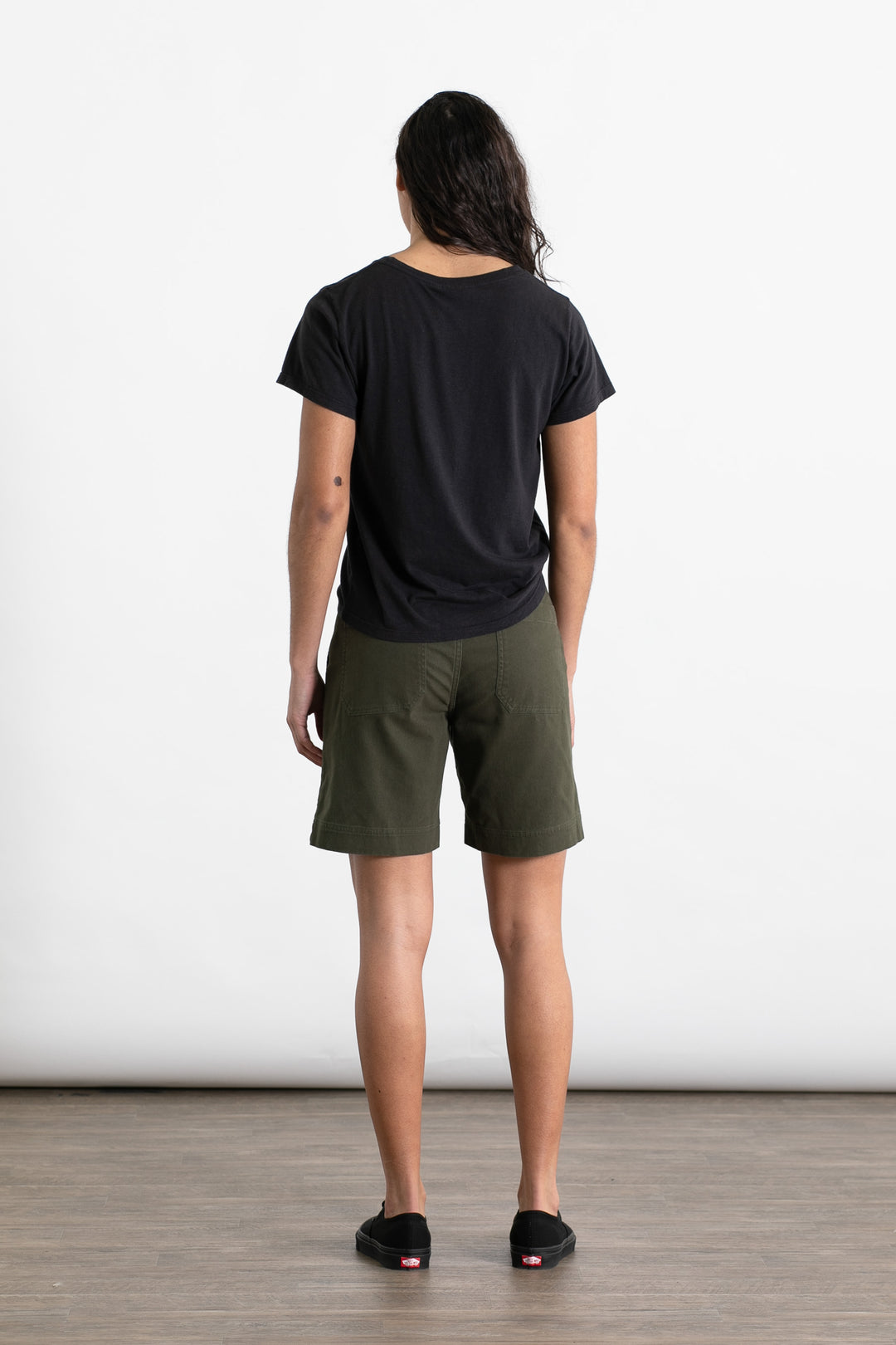 A person with long dark hair stands facing away on a wooden floor with a plain white background, wearing the Jungmaven Ojai Tee in black and olive green shorts paired with black slip-on shoes.