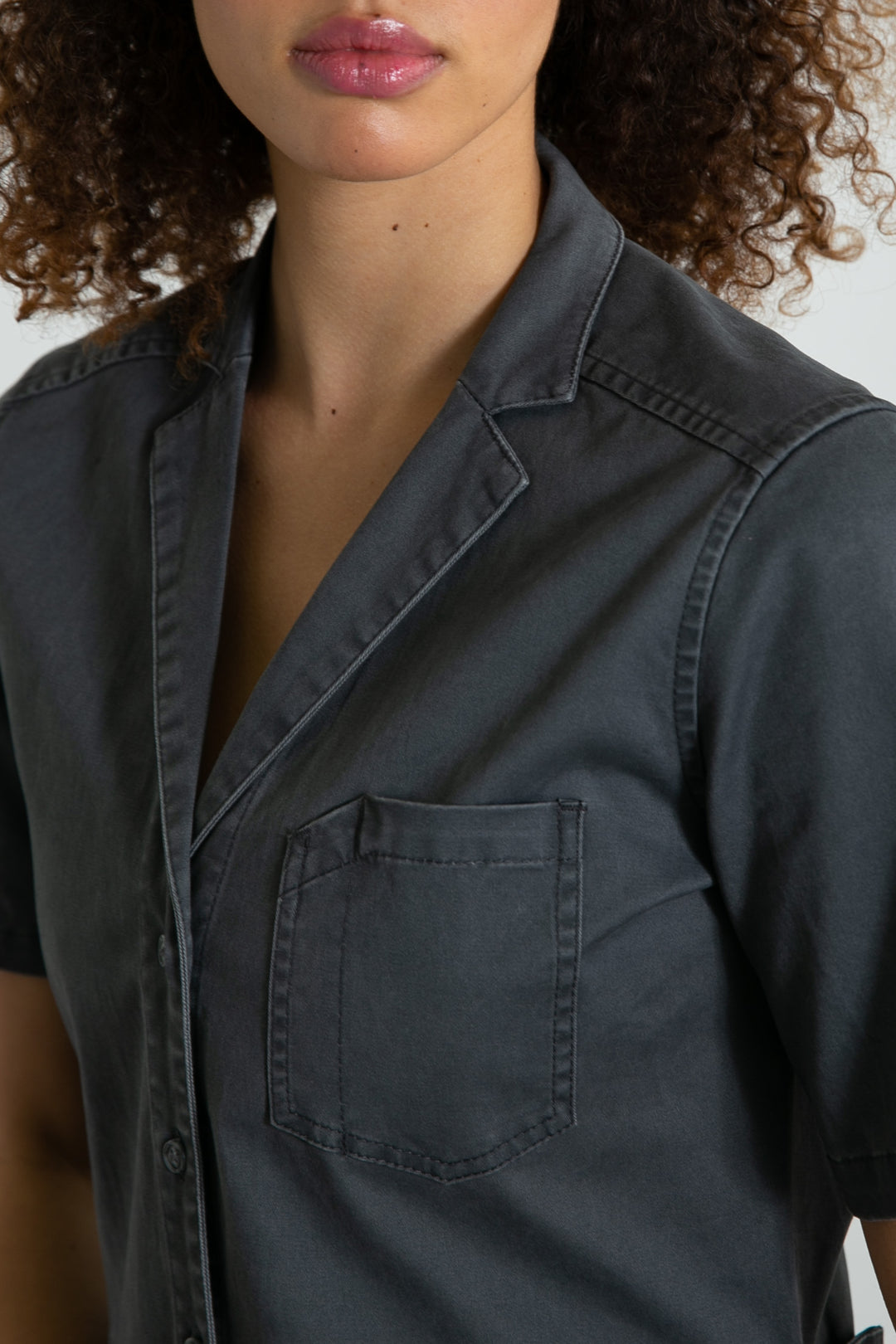 A woman with curly hair wears the Bridge & Burn Lou Utility Jumpsuit in Slate. The photo, showing her from shoulders to mid-torso with a partially visible face, highlights the short sleeves, chest pocket, and button-up design.