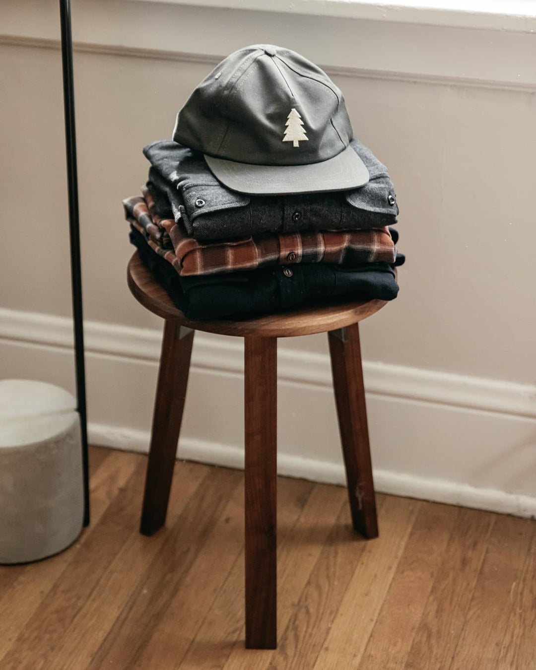 A stack of folded clothes topped with the Bridge & Burn Tree Cap in Olivewood, featuring a white tree logo, sits on a wooden stool in a room with wooden floors and white walls.